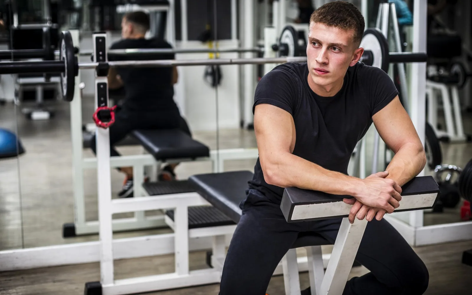 Hombre joven con camiseta negra sentado en un banco del gimnasio, apoyado en el respaldo mientras descansa, con una barra cargada con discos detrás.
